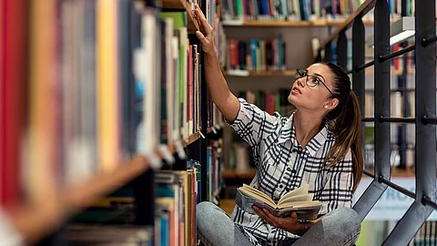 Eine junge Studentin sitzt in der Bücherei auf dem Boden zwischen Regalen und schaut nach Büchern.