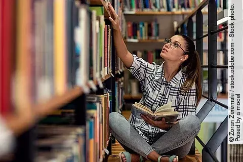 Eine junge Studentin sitzt in der Bücherei auf dem Boden zwischen Regalen und schaut nach Büchern.