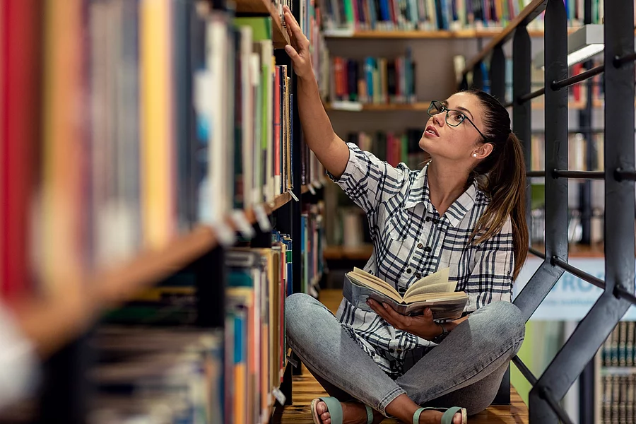 Eine junge Studentin sitzt in der Bücherei auf dem Boden zwischen Regalen und schaut nach Büchern.