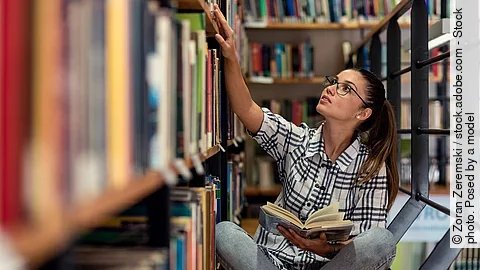 Eine junge Studentin sitzt in der Bücherei auf dem Boden zwischen Regalen und schaut nach Büchern.