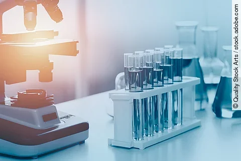 Wissenschaftliche medizinische Laborgeräte stehen auf einem Tisch mit einem Mikroskop im Hintergrund. Mehrere Reagenzgläser stehen in einer Halterung vor dem Mikroskop. Dahinter stehen befüllte Erlenmeierkolben. 