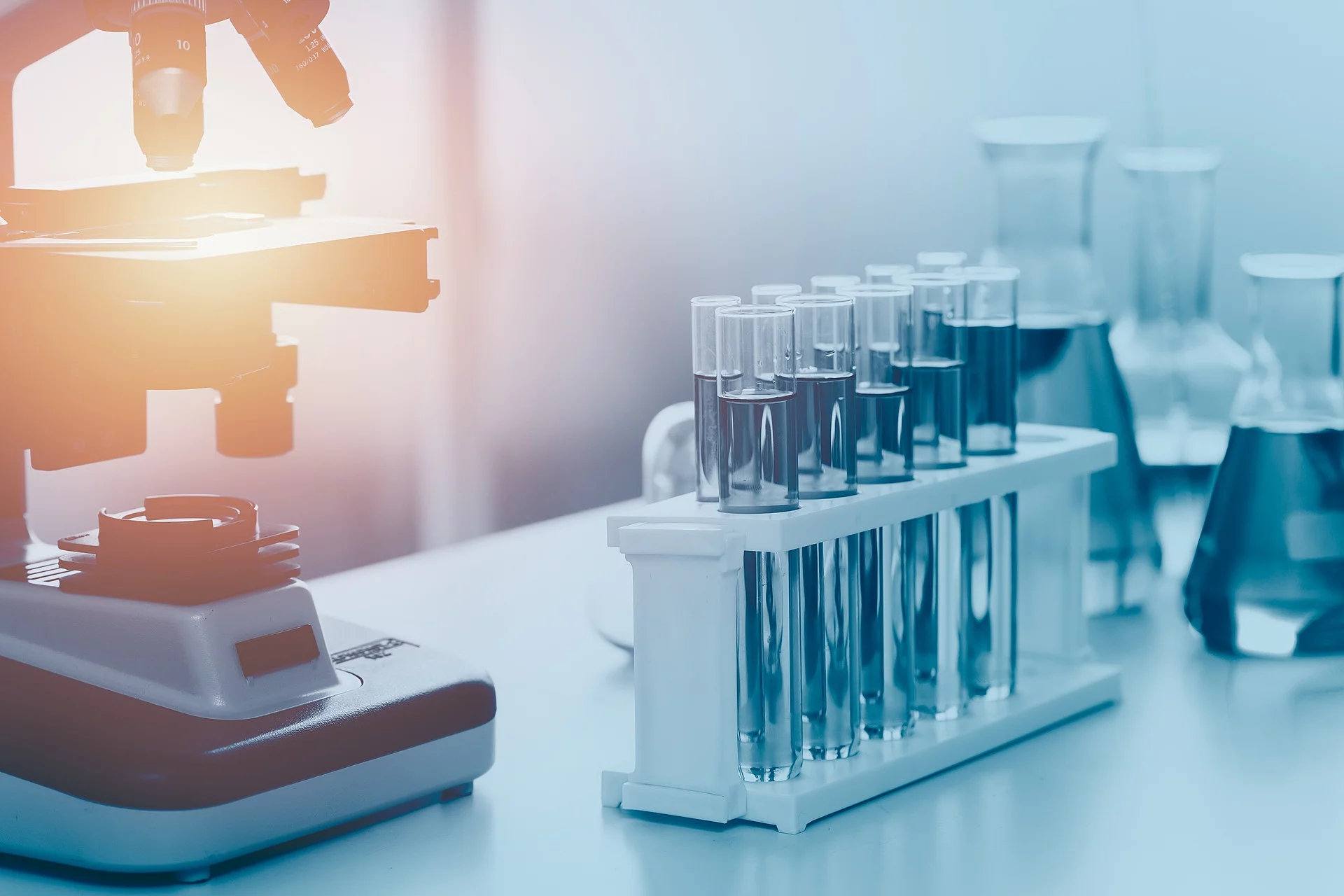 Wissenschaftliche medizinische Laborgeräte stehen auf einem Tisch mit einem Mikroskop im Hintergrund. Mehrere Reagenzgläser stehen in einer Halterung vor dem Mikroskop. Dahinter stehen befüllte Erlenmeierkolben. 