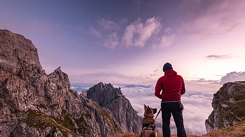 Ein Mann steht mit seinem Schäferhund zusammen auf einem Berggipfel und genießt die Aussicht. Der Himmel leuchtet in allen Rosatönen, da gerade die Sonne aufgeht.