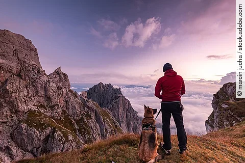 Ein Mann steht mit seinem Schäferhund zusammen auf einem Berggipfel und genießt die Aussicht. Der Himmel leuchtet in allen Rosatönen, da gerade die Sonne aufgeht.