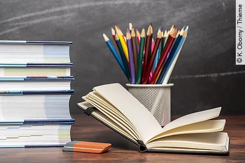 Ein aufgeschlagenes Buch liegt neben weiteren Büchern und Stiften auf einem Schreibtisch.