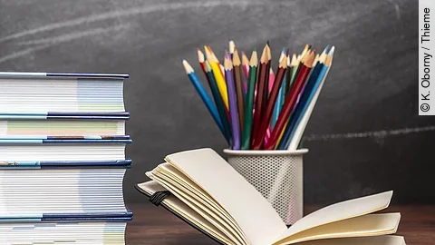 Ein aufgeschlagenes Buch liegt neben weiteren Büchern und Stiften auf einem Schreibtisch.