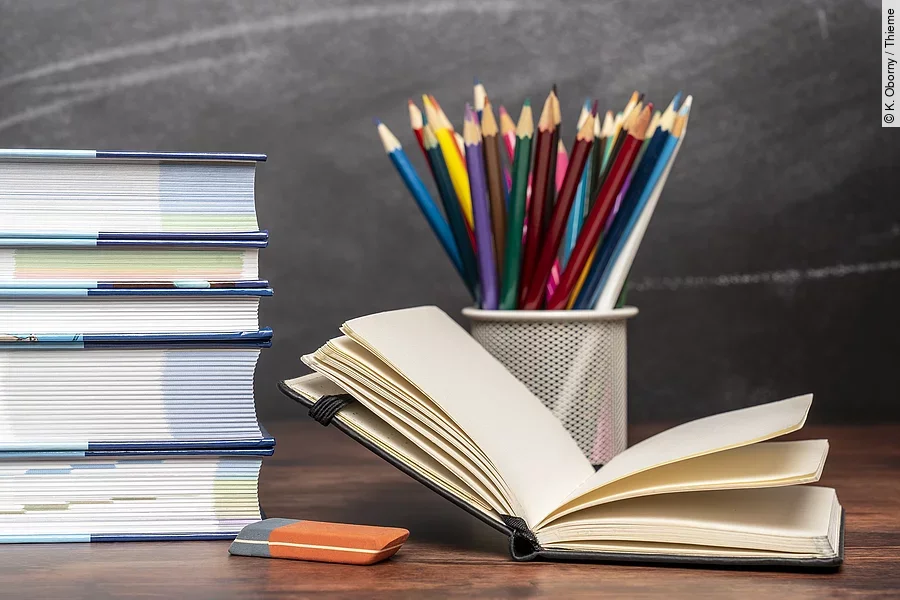 Ein aufgeschlagenes Buch liegt neben weiteren Büchern und Stiften auf einem Schreibtisch.