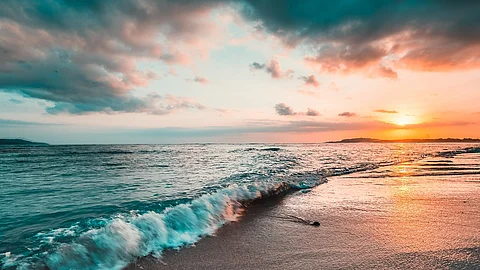 Das Meer von der Küste aus fotografiert. Im Hintergrund sieht man einen bunten Himmel durch die Sonne.