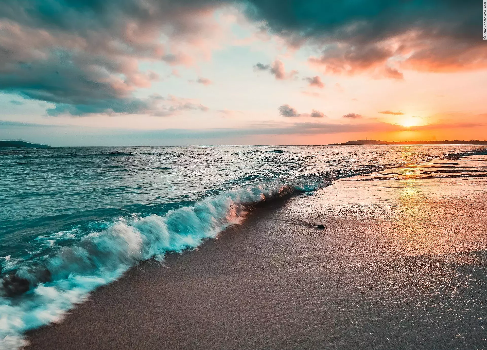 Das Meer von der Küste aus fotografiert. Im Hintergrund sieht man einen bunten Himmel durch die Sonne.