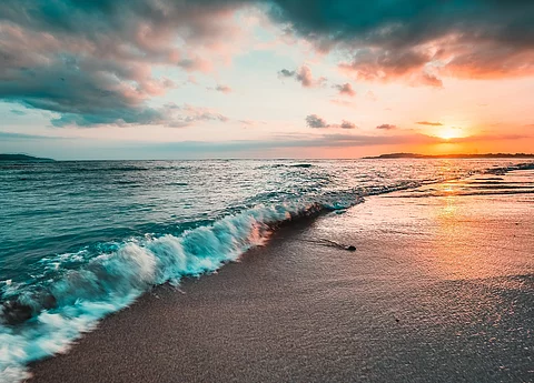 Das Meer von der Küste aus fotografiert. Im Hintergrund sieht man einen bunten Himmel durch die Sonne.