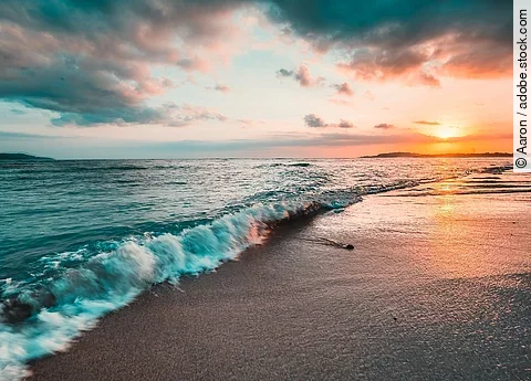 Das Meer von der Küste aus fotografiert. Im Hintergrund sieht man einen bunten Himmel durch die Sonne.