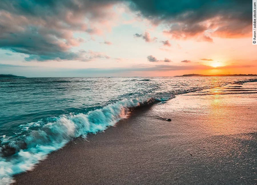 Das Meer von der Küste aus fotografiert. Im Hintergrund sieht man einen bunten Himmel durch die Sonne.