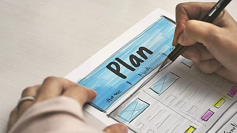 Ein Planzettel liegt im Fokus des Bildes, darauf schreibt eine Person mit Stift..