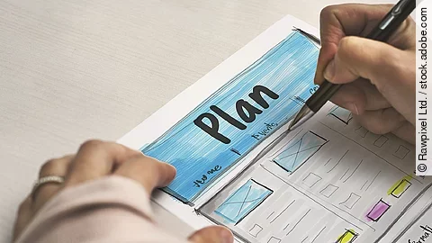 Ein Planzettel liegt im Fokus des Bildes, darauf schreibt eine Person mit Stift..