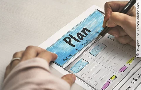 Ein Planzettel liegt im Fokus des Bildes, darauf schreibt eine Person mit Stift..