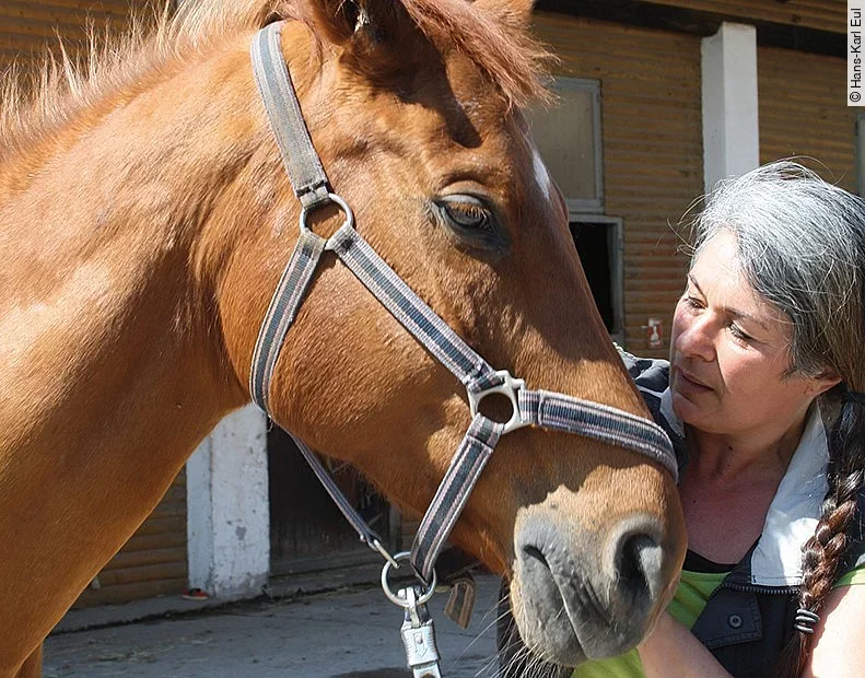 Frau Eul-Matern steht neben einem Pferd. Das Pferd trägt ein Halfter und hat die Augen geschlossen. Sein Fell ist fuchsfarben. 
