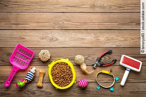 Auf dem Fußboden liegen unterschiedliche Haustier-Bedarfsartikel. Darunter eine pinke Katzenstreuschaufel, eine gelbe Futterschale voll mit Trockenfutter, verschiedenes Spielzeug und eine Bürste.