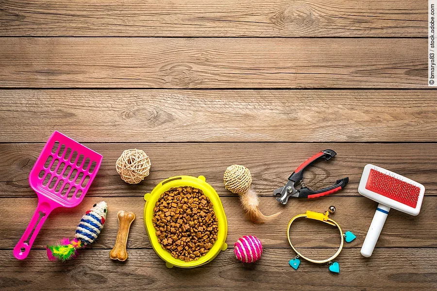 Auf dem Fußboden liegen unterschiedliche Haustier-Bedarfsartikel. Darunter eine pinke Katzenstreuschaufel, eine gelbe Futterschale voll mit Trockenfutter, verschiedenes Spielzeug und eine Bürste.