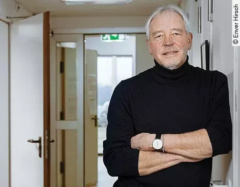 Professor Thomas Bosch trägt einen schwarzen Pullover und eine schwarze Hose. Vor seinem Körper hat er seine Arme verschränkt und er lehnt lässig gegen eine Wand.