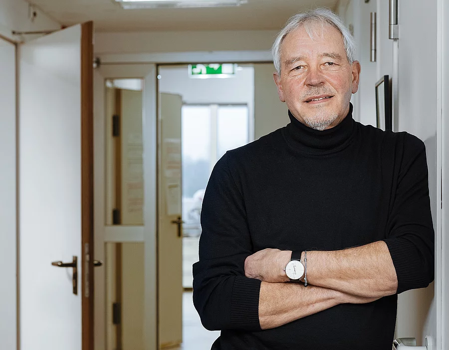 Professor Thomas Bosch trägt einen schwarzen Pullover und eine schwarze Hose. Vor seinem Körper hat er seine Arme verschränkt und er lehnt lässig gegen eine Wand.
