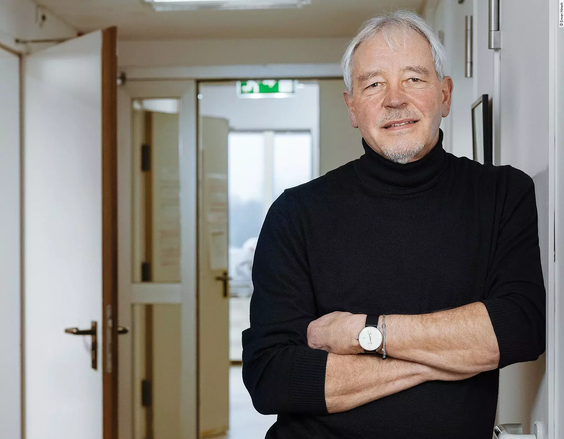 Professor Thomas Bosch trägt einen schwarzen Pullover und eine schwarze Hose. Vor seinem Körper hat er seine Arme verschränkt und er lehnt lässig gegen eine Wand.