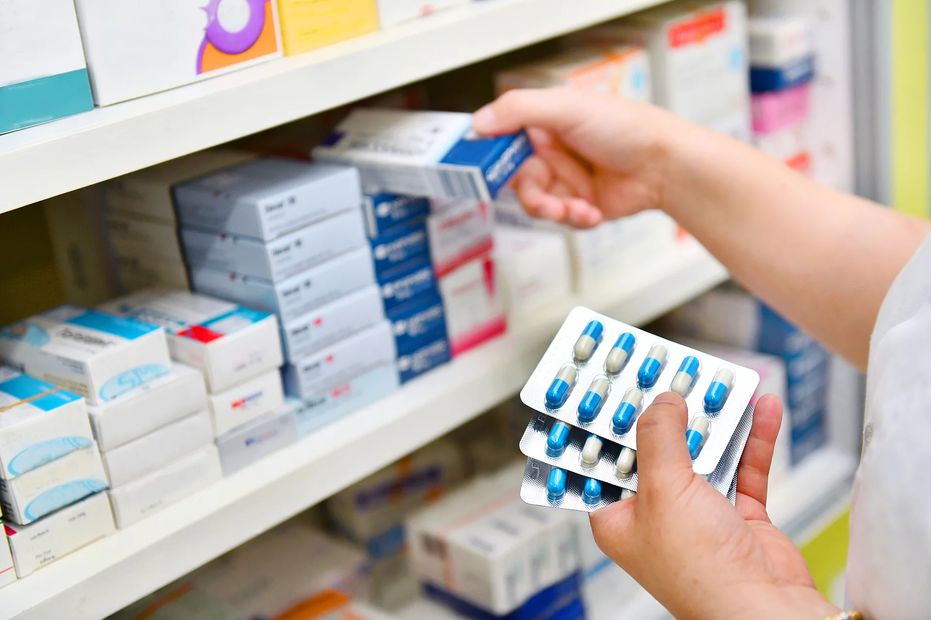Ein Apotheker räumt Arzneimittel in ein Regal ein. Dabei hält er mehrere Tablettenblister in der einen Hand und eine Arzneimittelpackung in der anderen.