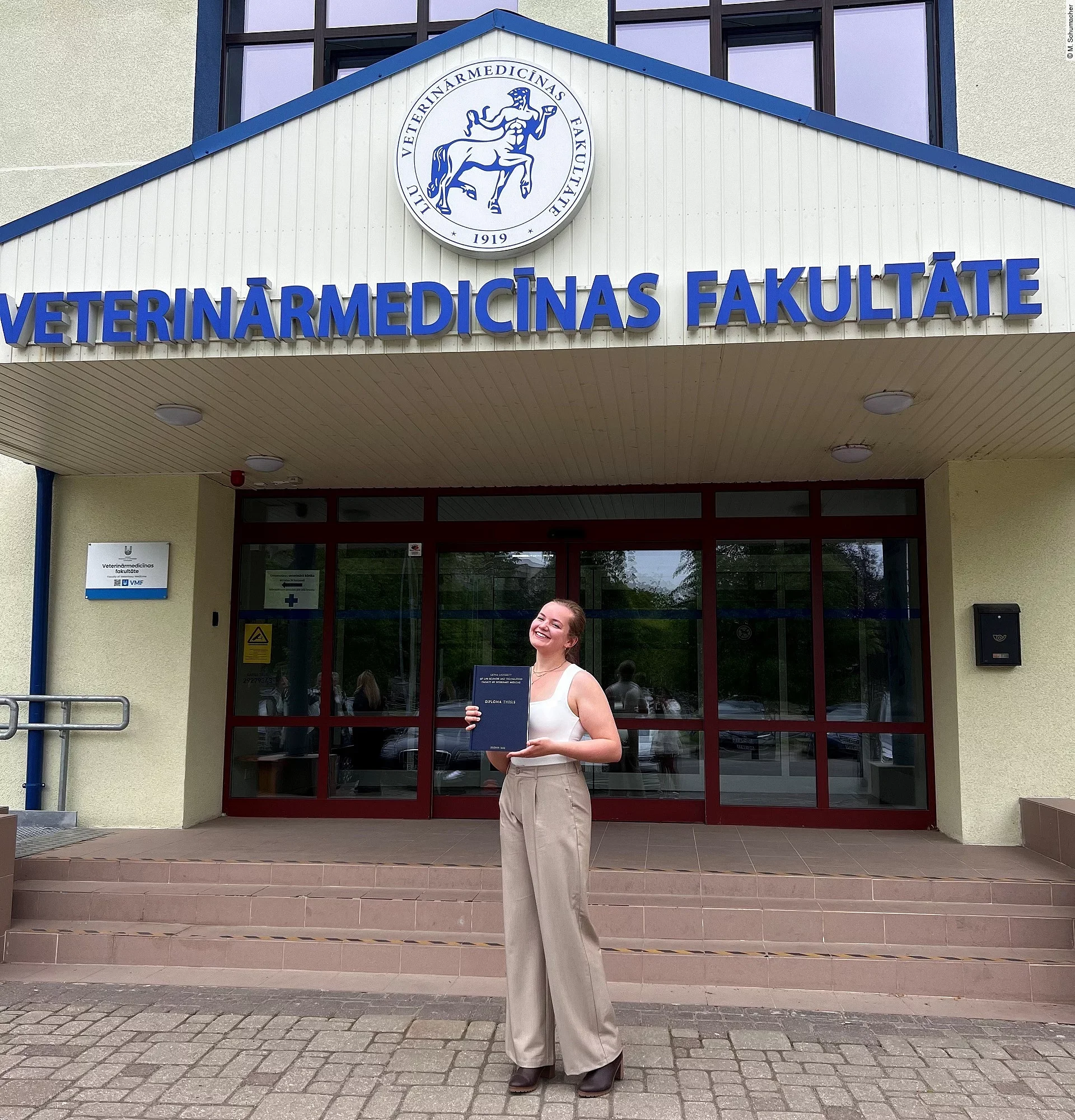 Merle steht mit ihrer Diplomarbeit in der Hand vor dem Universitätsgebäude. Sie trägt schicke Kleidung und lächelt in die Kamera.