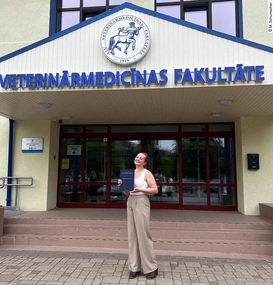 Merle steht mit ihrer Diplomarbeit in der Hand vor dem Universitätsgebäude. Sie trägt schicke Kleidung und lächelt in die Kamera.