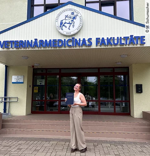 Merle steht mit ihrer Diplomarbeit in der Hand vor dem Universitätsgebäude. Sie trägt schicke Kleidung und lächelt in die Kamera.