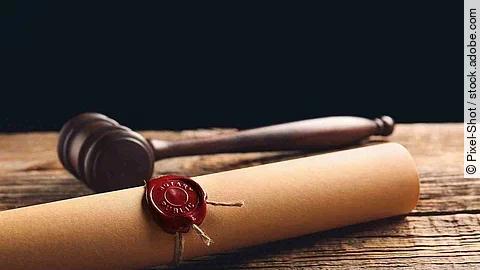 Eine Papierrolle mit rotem Siegel liegt auf einem Holztisch. Daneben liegt ein hölzerner Richterhammer.