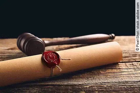 Eine Papierrolle mit rotem Siegel liegt auf einem Holztisch. Daneben liegt ein hölzerner Richterhammer.