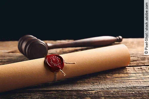 Eine Papierrolle mit rotem Siegel liegt auf einem Holztisch. Daneben liegt ein hölzerner Richterhammer.