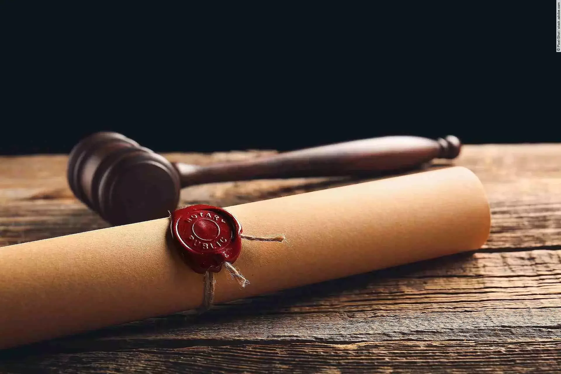 Eine Papierrolle mit rotem Siegel liegt auf einem Holztisch. Daneben liegt ein hölzerner Richterhammer.