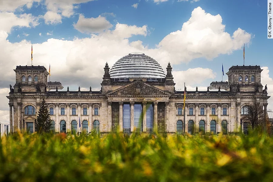 Aufnahme vom Reichstagsgebäude in Berlin. Das Gebäude ist von unten fotografiert und im Hintergrund ist ein blauer Himmel mit Wolken. Im Vordergrund des Bildes ist eine grüne Wiese.