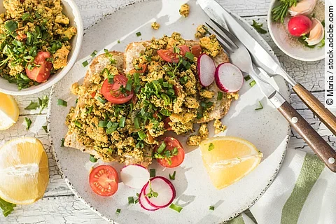 Aufnahme von einem knusprigen Toastbrot mit Rührei aus Tofu und Tomatensalat. Über dem Tofu ist Schnittlauch gestreut. Das Essen ist auf einem Teller angerichtet und daneben liegen ein Set Messer und Gabel.