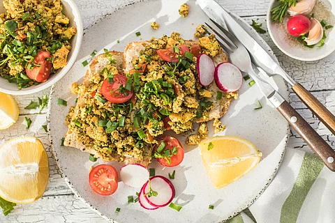 Aufnahme von einem knusprigen Toastbrot mit Rührei aus Tofu und Tomatensalat. Über dem Tofu ist Schnittlauch gestreut. Das Essen ist auf einem Teller angerichtet und daneben liegen ein Set Messer und Gabel.