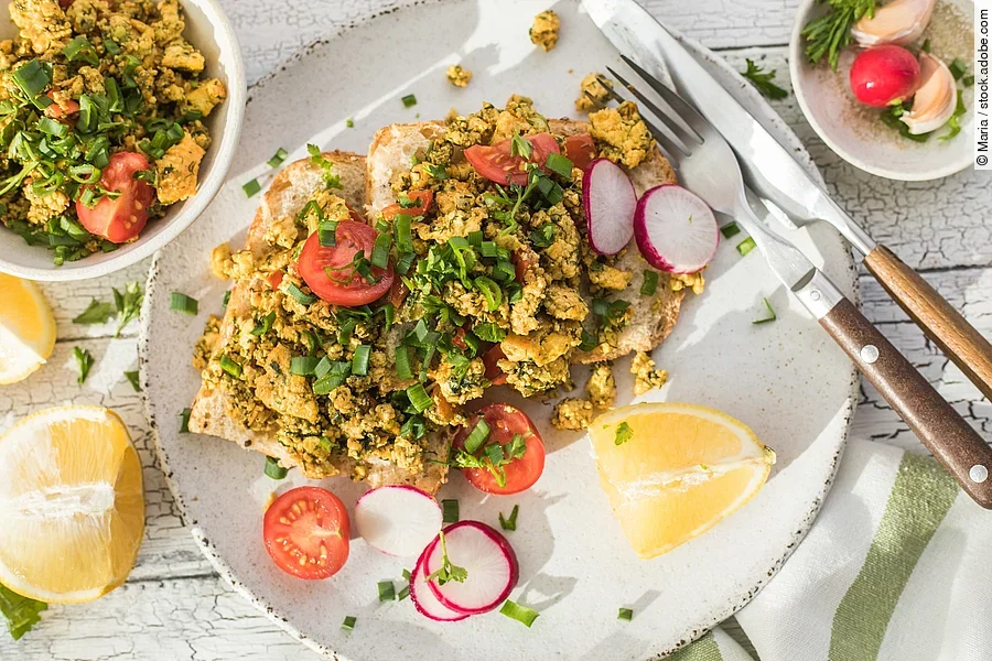 Aufnahme von einem knusprigen Toastbrot mit Rührei aus Tofu und Tomatensalat. Über dem Tofu ist Schnittlauch gestreut. Das Essen ist auf einem Teller angerichtet und daneben liegen ein Set Messer und Gabel.