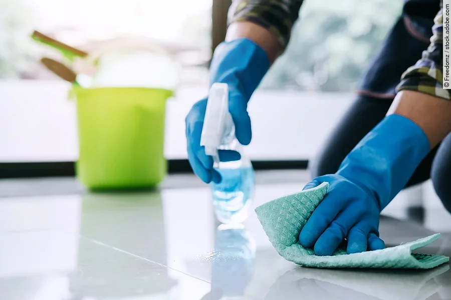 Eine Person wischt mit einem Lappen über dem Boden. In der zweiten Hand wird eine Sprühflasche gehalten,