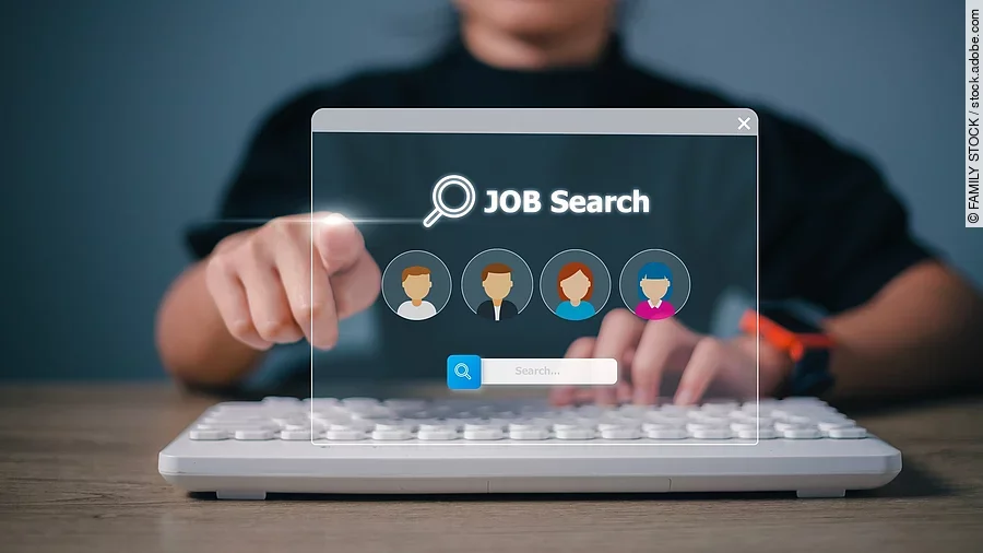 Ein Person sitzt an einem braunen Holztisch und vor ihr liegt eine weiße Tastatur. Ihre linke Hand liegt auf der Tastatur und die rechte Hand zeigt mit einem Zeigefinger nach vorne. Vor der Person ist eine digitale Jobsuche als Hologramm. Auf die Jobsuche zeigt der Zeigefinger. Die Person trägt ein schwarzes Oberteil und eine Smartwacht am Handgelenk. Die Person ist nicht identifizierbar, da das Bild nur bis zu den Schultern reicht. 