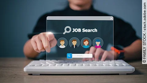 Ein Person sitzt an einem braunen Holztisch und vor ihr liegt eine weiße Tastatur. Ihre linke Hand liegt auf der Tastatur und die rechte Hand zeigt mit einem Zeigefinger nach vorne. Vor der Person ist eine digitale Jobsuche als Hologramm. Auf die Jobsuche zeigt der Zeigefinger. Die Person trägt ein schwarzes Oberteil und eine Smartwacht am Handgelenk. Die Person ist nicht identifizierbar, da das Bild nur bis zu den Schultern reicht. 