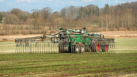 Ein Traktor bring Gülle auf das Feld aus. Der Trecker ist groß und verbringt per Schleppschlauch Gülle auf das Feld. Auf dem Feld sprießen kurze grüne Pflanzen und im Hintergrund stehen Bäume. 