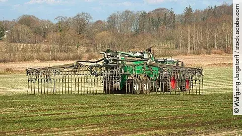 Ein Traktor bring Gülle auf das Feld aus. Der Trecker ist groß und verbringt per Schleppschlauch Gülle auf das Feld. Auf dem Feld sprießen kurze grüne Pflanzen und im Hintergrund stehen Bäume. 