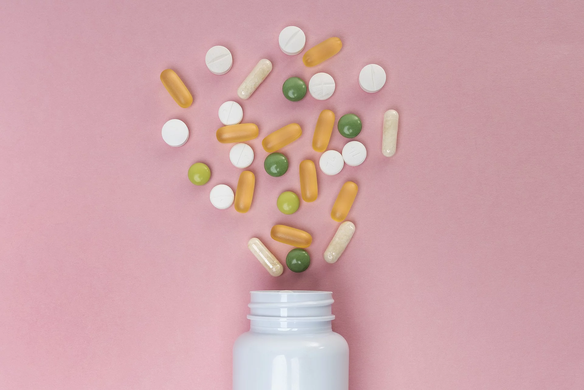 Aus einer Pillendose kommen grüne, weiße und gelbe Tabletten und Kapseln raus. Dabei liegt alles auf einem rosafarbenen Untergrund.