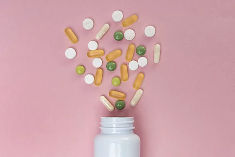 Aus einer Pillendose kommen grüne, weiße und gelbe Tabletten und Kapseln raus. Dabei liegt alles auf einem rosafarbenen Untergrund.