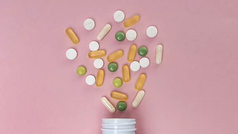 Aus einer Pillendose kommen grüne, weiße und gelbe Tabletten und Kapseln raus. Dabei liegt alles auf einem rosafarbenen Untergrund.