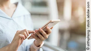 Eine Frau nutzt ihr Smartphone. Die Person trägt eine Bluse. Das Handy ist ohne Hülle. Die Person trägt eine Armbanduhr. Der Hintergrund ist verschwommen.