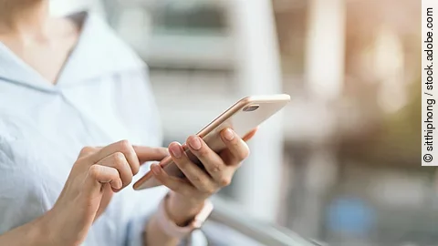 Eine Frau nutzt ihr Smartphone. Die Person trägt eine Bluse. Das Handy ist ohne Hülle. Die Person trägt eine Armbanduhr. Der Hintergrund ist verschwommen.