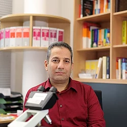 Professor Hamedy sitzt in einem Büro mit großer Bücherwand vor einem Mikroskop.