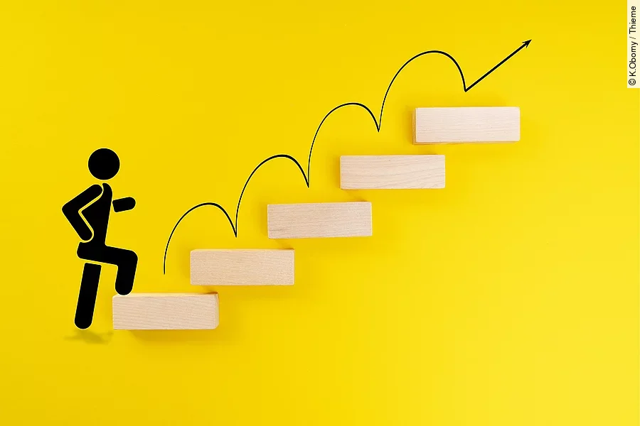 Ein schwarzes Ampelmännchen läuft eine Treppe hinauf. Die einzelnen Stufen bestehen aus braunen Holzbausteinen und liegen vor einem gelben Hintergrund.