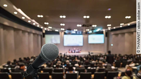 Ein Mikrofon steht am Ende einer großen Kongresshalle. Auf den Stühlen davor sitzen viele Teilnehmende und schauen gespannt auf die Leinwand.